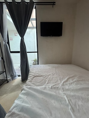 Una habitación con cama, cortinas, televisor montado en la pared y ventana con vistas al exterior.