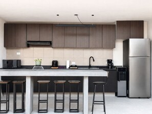 A modern kitchen with dark wooden cabinets, a stainless steel refrigerator, a built-in microwave, a coffee maker, and a bar stool area. - Hotel Casa Rosa (Aguascalientes)