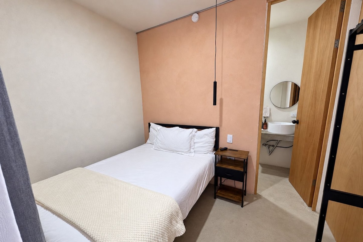 A bedroom with a single bed, a bedside table, a nightstand, a wooden wardrobe, and a bathroom visible through an open door.