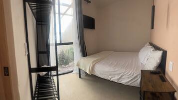 Una habitación con cama, una mesa de madera, televisor montado en la pared y ventana con cortinas.