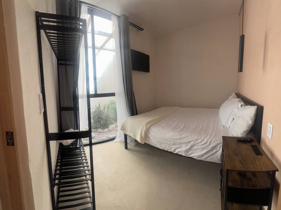 Una habitación con cama, una mesa de madera, televisor montado en la pared y ventana con cortinas.
