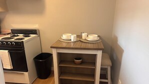 Una cocina compacta con estufa, un bote de basura, una pequeña mesa con tazas y fuentes, y una silla.