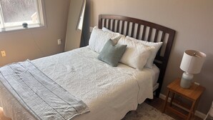 Una cama bien hecha con una colcha y almohadas estampadas, una mesita de noche con una lámpara y vistas al exterior a través de una ventana.