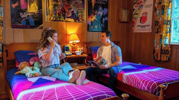 A room with two wooden beds, posters on the walls, a window with curtains, and a lamp on a nightstand.