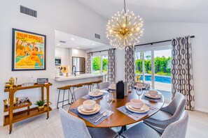 A dining area with a table set for six, a modern chandelier, and a view of a swimming pool. - Sun and Games - Stylish Desert Retreat (Palm Springs)
