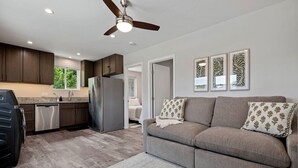 A modern kitchen with dark cabinets, a stainless steel refrigerator, and a dishwasher. There is a gray sofa with patterned pillows and a ceiling fan.