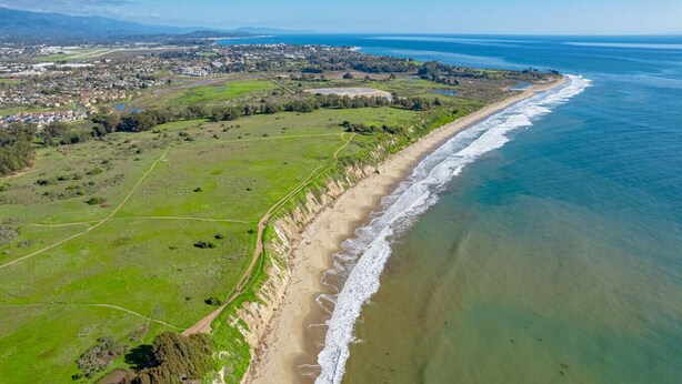 An aerial view of a coastal area with a beach, green fields, and a distant town.