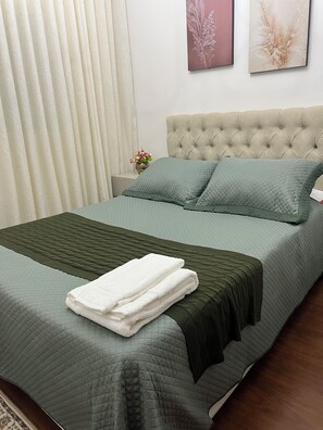 Ein ordentlich bezogenes Bett mit einer gesteppten, grünen Tagesdecke und Kissen, ein weißes Handtuch auf dem Bett und ein gebürstetes Kopfstück.