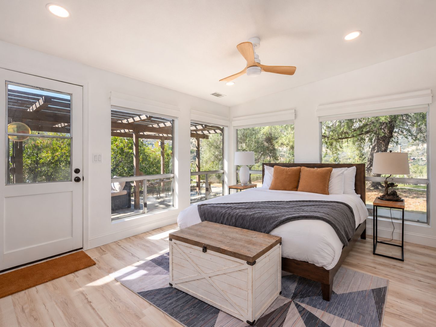 A bedroom with a bed, a nightstand, a lamp, a ceiling fan, and large windows.