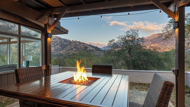 Un patio al aire libre con una mesa de madera, dos sillas de mimbre, una chimenea y vistas a montañas.