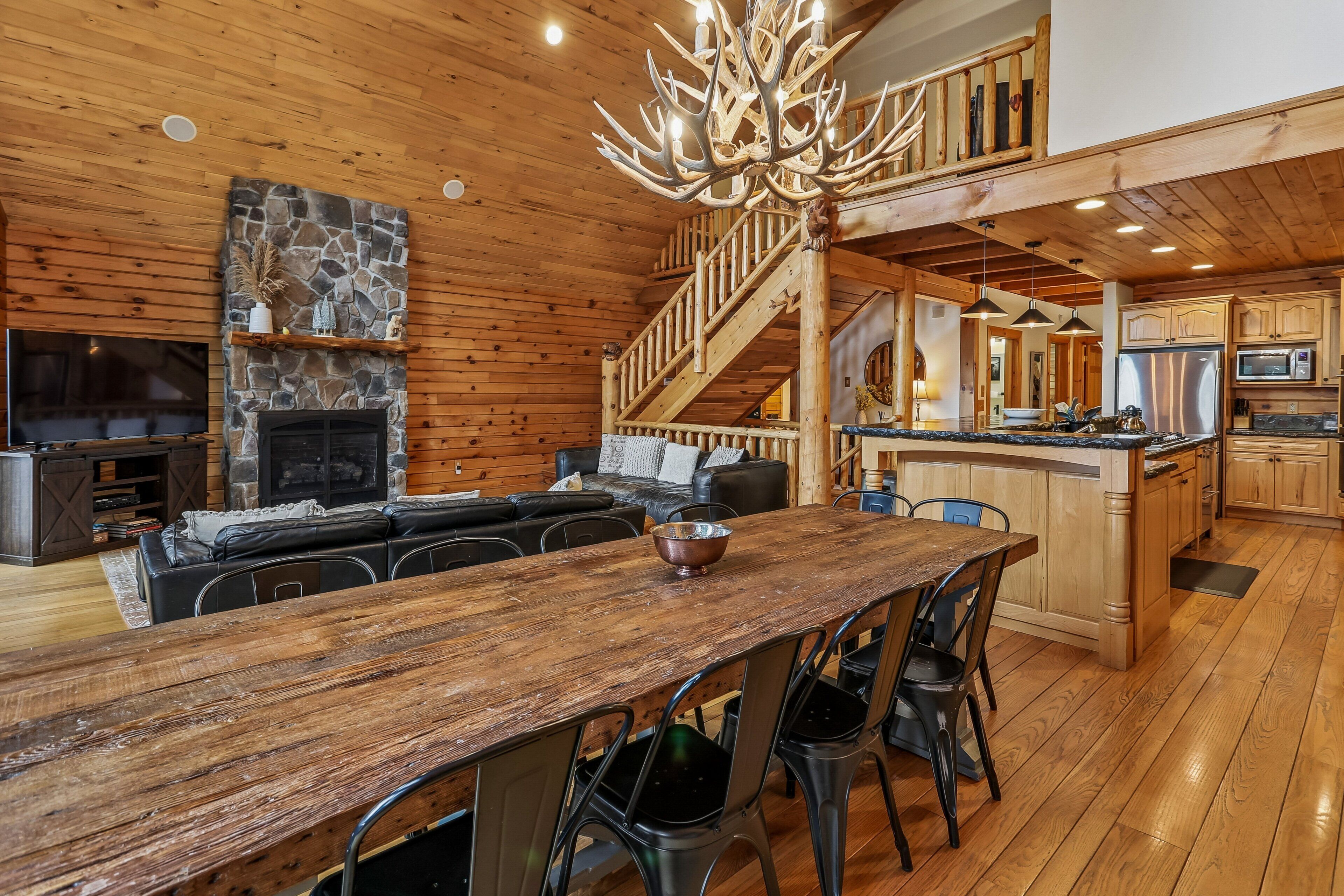 Un intérieur rustique avec une cheminée en pierre, un plafond en bois, un coin repas avec une longue table et une cuisine équipée d’appareils modernes.
