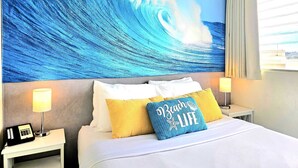 Et hotelværelse med et stort maleri af en bølge på væggen, en seng med hvidt sengetøj og en pude med teksten 'Beach Life'.