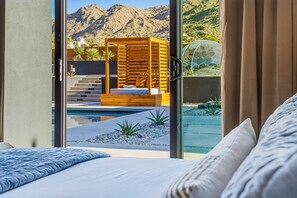 A bedroom with a large window, a bed, and a view of a pool and mountains. - Skyhaven Luxe - Pool/Spa Pickleball Sauna (Joshua Tree)