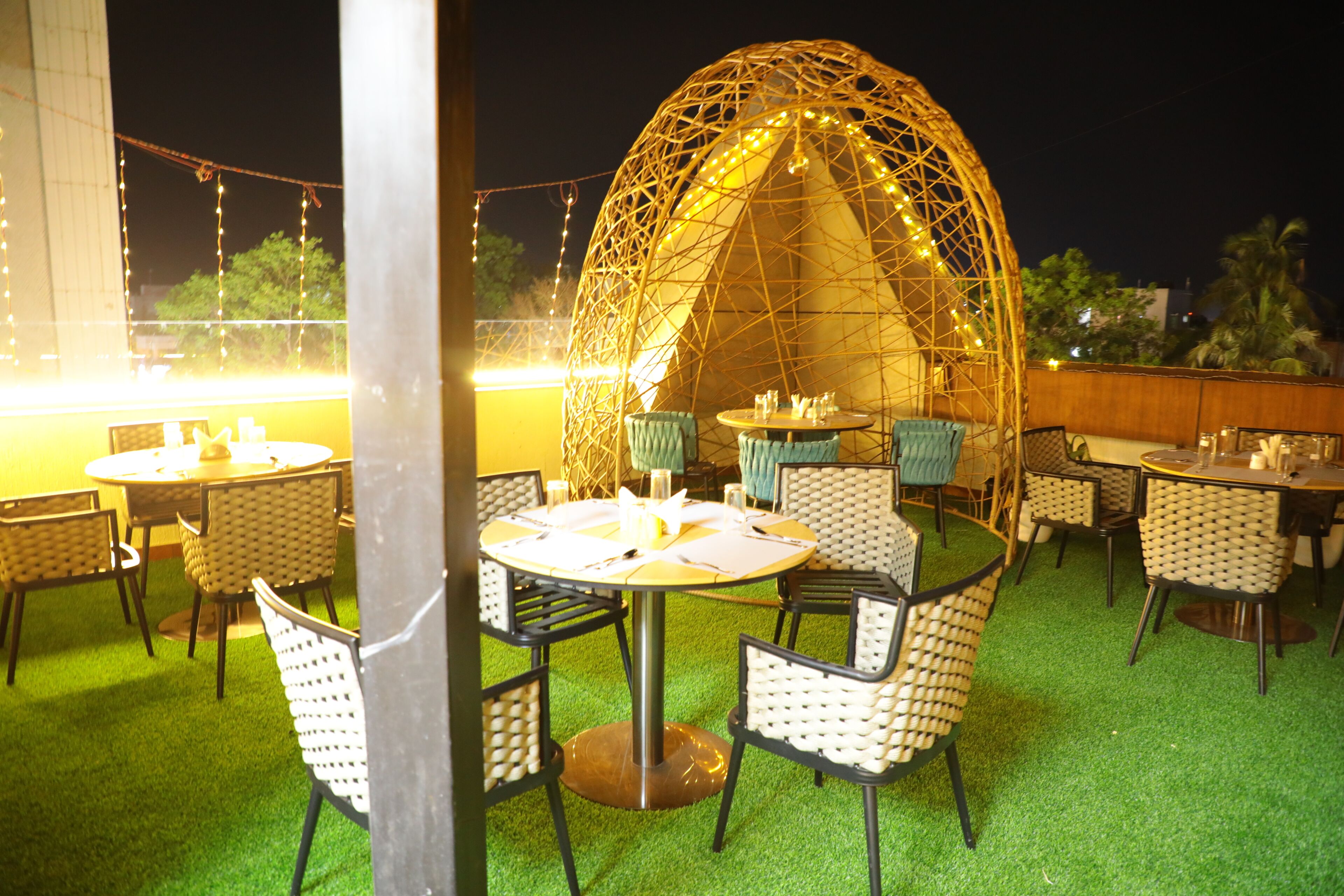 An outdoor dining area with round tables and chairs, a decorative arch structure, and string lights.