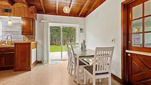 A kitchen with wooden cabinets, a dining table with chairs, and a view through a glass door to a garden. - 2BR Coastal Cottage Near Downtown & Beaches (Vero Beach)
