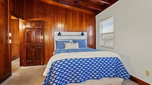 A bedroom with a wooden door, a bed with a blue and white patterned bedspread, a white headboard, and a window with blinds. - 2BR Coastal Cottage Near Downtown & Beaches (Vero Beach)