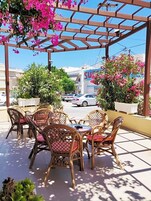 Un área de estar al aire libre con sillas y mesas de mimbre, rodeada de macetas con plantas y flores.