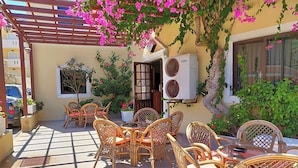 Un patio al aire libre con mobiliario de mimbre, una pérgola con flores rosadas y un equipo de aire acondicionado.