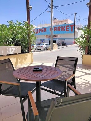 Et udendørs opholdsområde med et rundt bord og stole, en potteplante og udsigt til en gade med parkerede biler og en bygning med skiltet 'Super Market'.