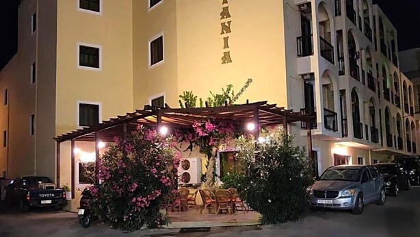 Un hotel con un cartel que dice 'HOTEL IANIA', con un espacio de estar al aire libre cubierto, con sillas y mesas, y un estacionamiento con coches.