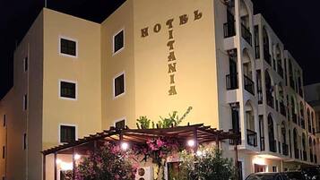 A hotel with a sign reading 'HOTEL IANIA,' featuring a covered outdoor seating area with chairs and tables, and a parking lot with cars.