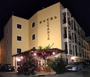 A hotel with a sign reading 'HOTEL IANIA,' featuring a covered outdoor seating area with chairs and tables, and a parking lot with cars. - Titania - Karpathos (Karpathos)