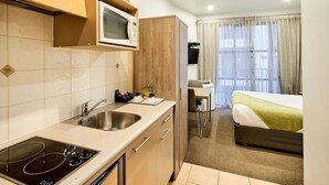 A compact hotel room with a kitchenette, a bed, a desk, and a window with curtains.