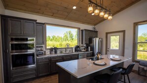 A modern kitchen with dark cabinets, stainless steel appliances, a large island with bar stools, and large windows offering a view of greenery. - Hazel Hollow | Hot Tub w/ TV • Steam Shower • RRG (Rogers)