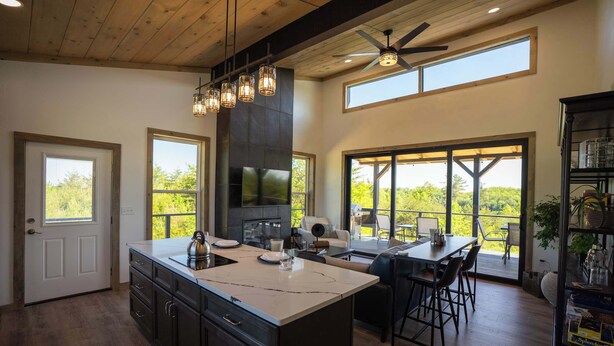 A modern kitchen with a large island, a dining area with a table and chairs, a fireplace, and large windows offering a view of the outdoors. - Hazel Hollow | Hot Tub w/ TV • Steam Shower • RRG (Rogers)