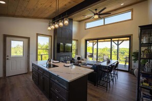 A modern kitchen with a large island, a dining area with a table and chairs, a fireplace, and large windows offering a view of the outdoors. - Hazel Hollow | Hot Tub w/ TV • Steam Shower • RRG (Rogers)