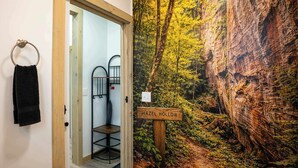 A bathroom with a wooden door, a towel ring, a metal chair, and a wall mural depicting a forest scene with a sign reading 'HAZEL HOLLOW.' - Hazel Hollow | Hot Tub w/ TV • Steam Shower • RRG (Rogers)