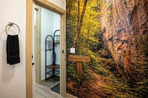 A bathroom with a wooden door, a towel ring, a metal chair, and a wall mural depicting a forest scene with a sign reading 'HAZEL HOLLOW.'