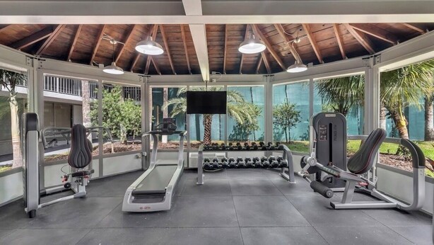 Ett gym med tak, utrustat med löpband, hantlar och styrketräningsmaskiner, stora fönster och en tv-skärm.