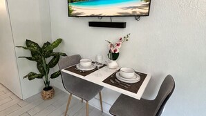En matplats med en tv monterad på väggen som visar en strandscen, ett bord dukat med tallrikar och glas, två stolar, en krukväxt och en liten vas med blommor.