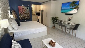 Ett modernt sovrum med en säng, en soffa, en matplats med stolar, en tv och ett kök i bakgrunden.