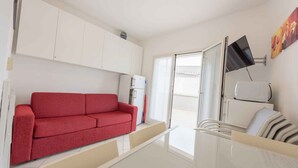 A modern living room with a red sofa, white cabinets, a flat-screen TV, and a sliding glass door leading to an outdoor area. - Holiday apartment with balcony and air conditioning (Bibione)