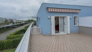 A blue building with a red and white awning, a gated balcony, and a view of a road with cars. - Holiday apartment with balcony and air conditioning (Bibione)