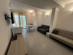 A living room with a sofa, a TV mounted on the wall, a dining table with chairs, and a small table with a box. - Holiday apartment with air conditioning and satellite TV (Bibione)