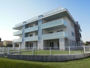 A multi-story building with balconies, a fenced courtyard, and a clear sky. - Holiday apartment with air conditioning and satellite TV (Bibione)