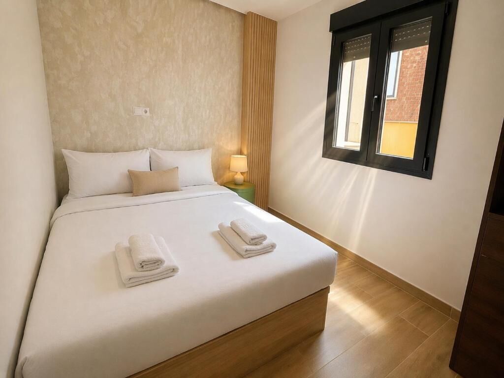 A neatly made bed with two folded towels, a bedside lamp, and a window with a view of a building.