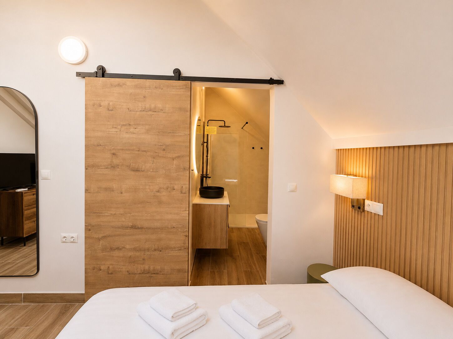 A hotel room with a bed, a wooden sliding door to a bathroom, a mirror, and a wall-mounted lamp.