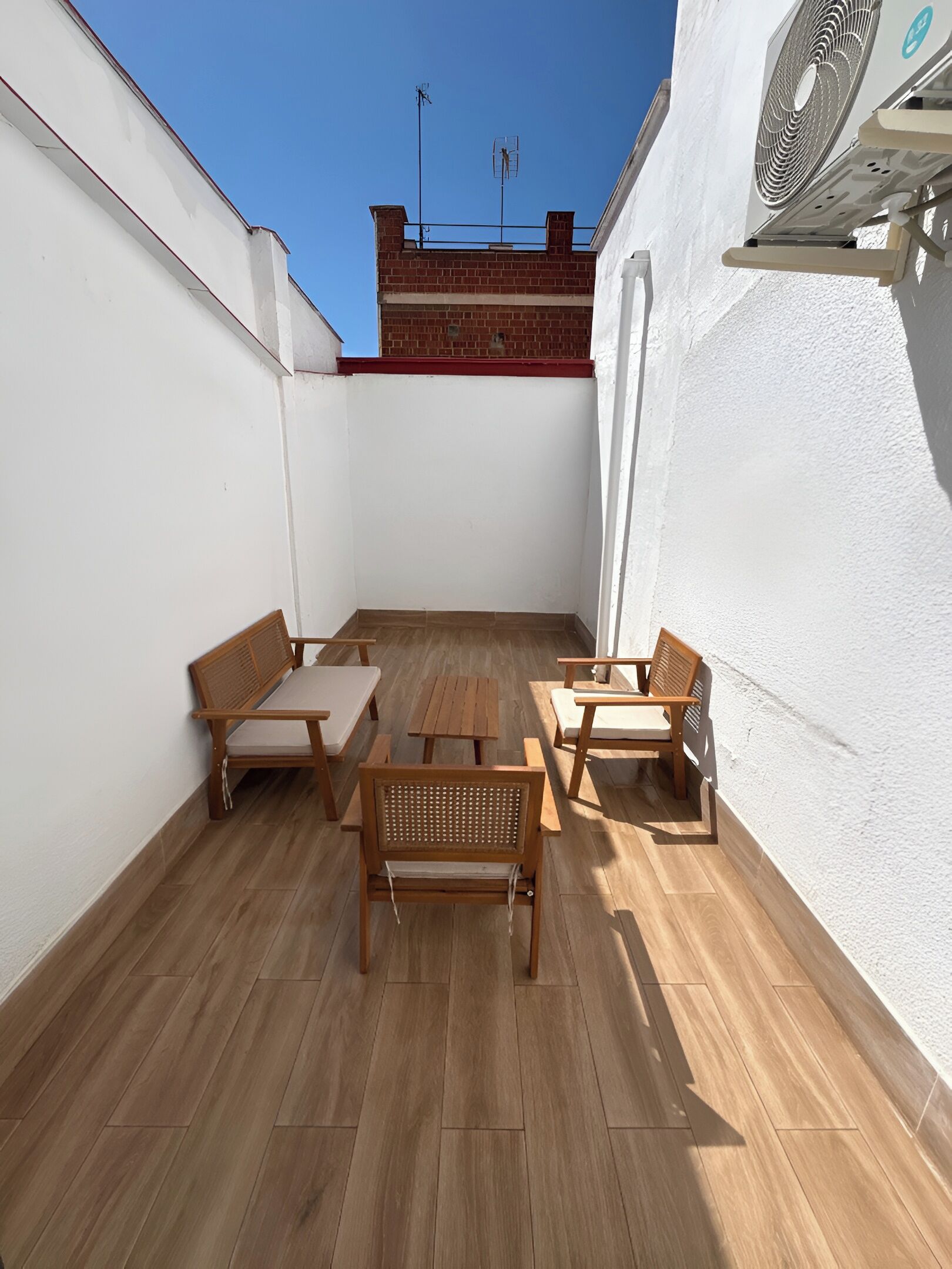 Un patio en la azotea con muebles de madera, un ventilador y un cielo azul despejado.