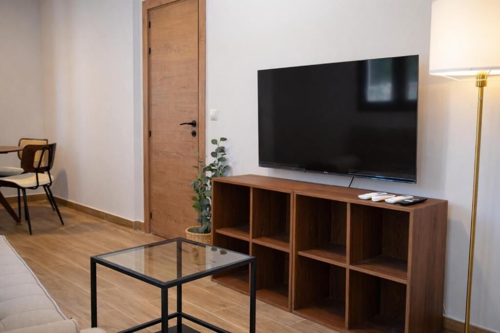 A living room with a flat-screen TV mounted on a wooden shelf, a glass coffee table, a sofa, a chair, and a floor lamp.