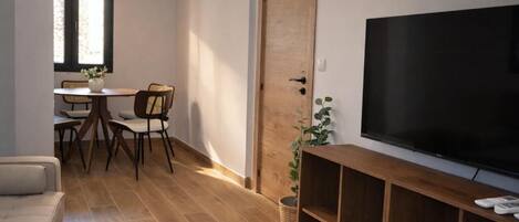 Una sala de estar con un sofá, una mesa de centro de vidrio, un televisor sobre un mueble de madera, una zona de comedor con sillas y una mesa, y una puerta de madera.