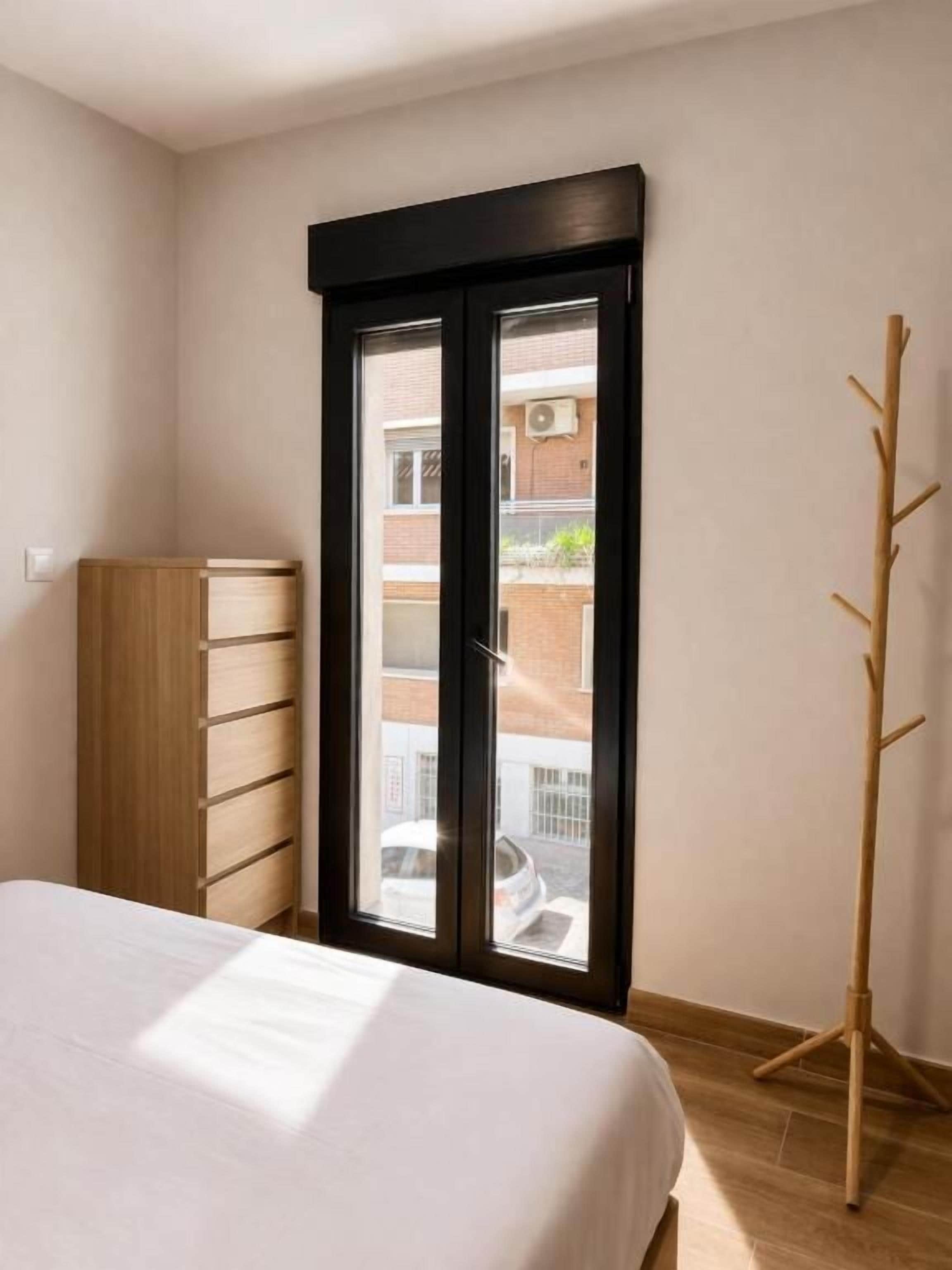 A bedroom with a bed, a wardrobe, a window with a view of a building, and a coat rack.