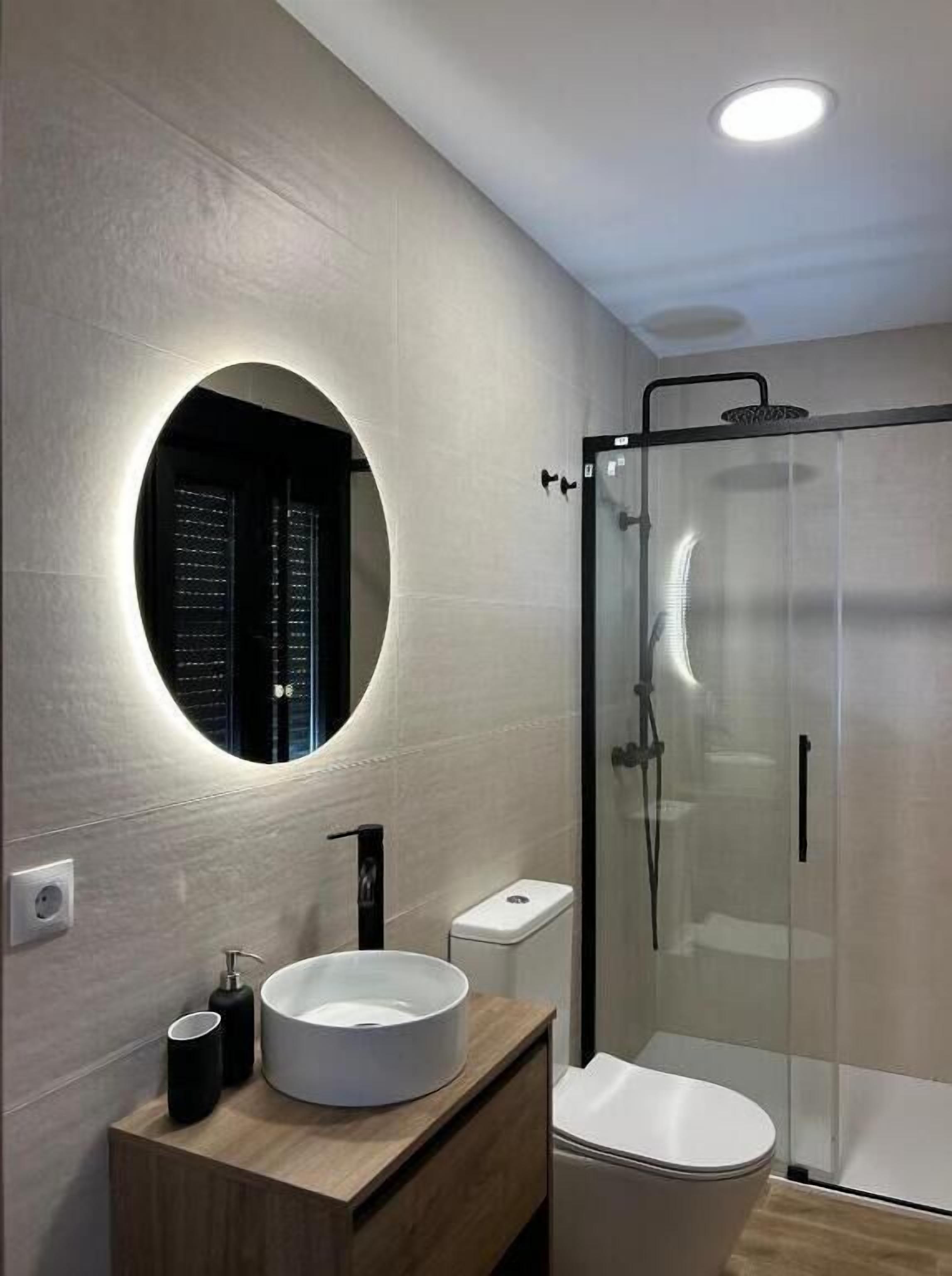 A modern bathroom with a round mirror, a sink, a toilet, and a glass-enclosed shower.