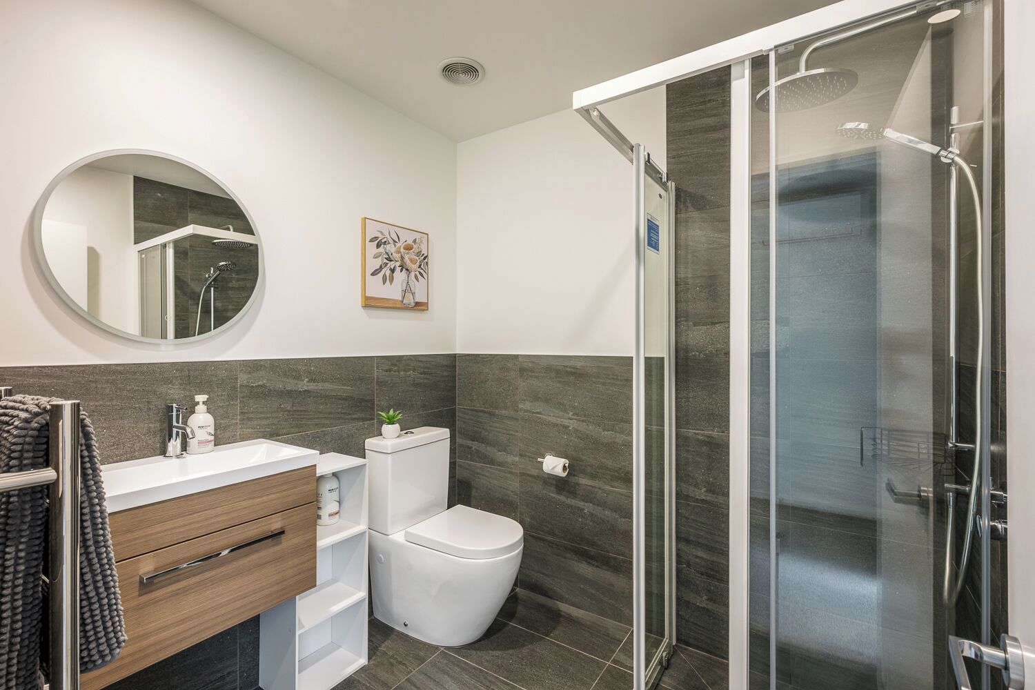 A modern bathroom with a glass shower enclosure, a round mirror, a toilet, and a sink with a wooden vanity.