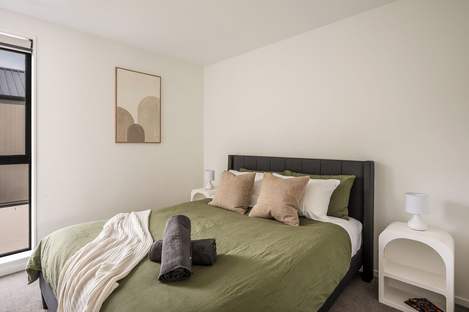 A bedroom with a bed, pillows, a blanket, a nightstand, a lamp, and a wall-mounted artwork.