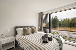 A bedroom with a bed, nightstand, lamp, and a view of a road and trees. - Relaxing Wānaka Townhouse (Wānaka)