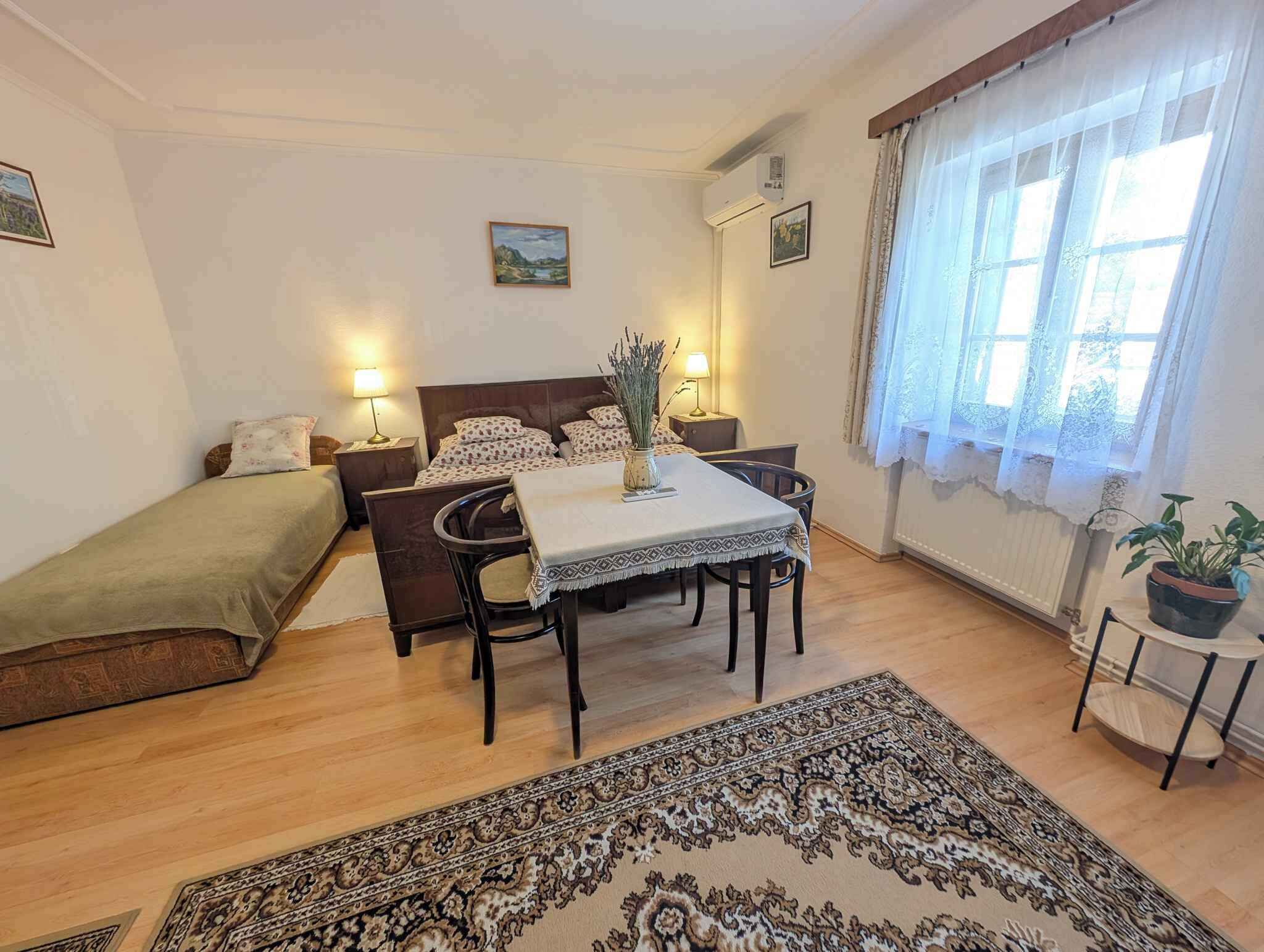 A bedroom with a bed, a small table with chairs, a bedside table with lamps, a window with curtains, and a potted plant.